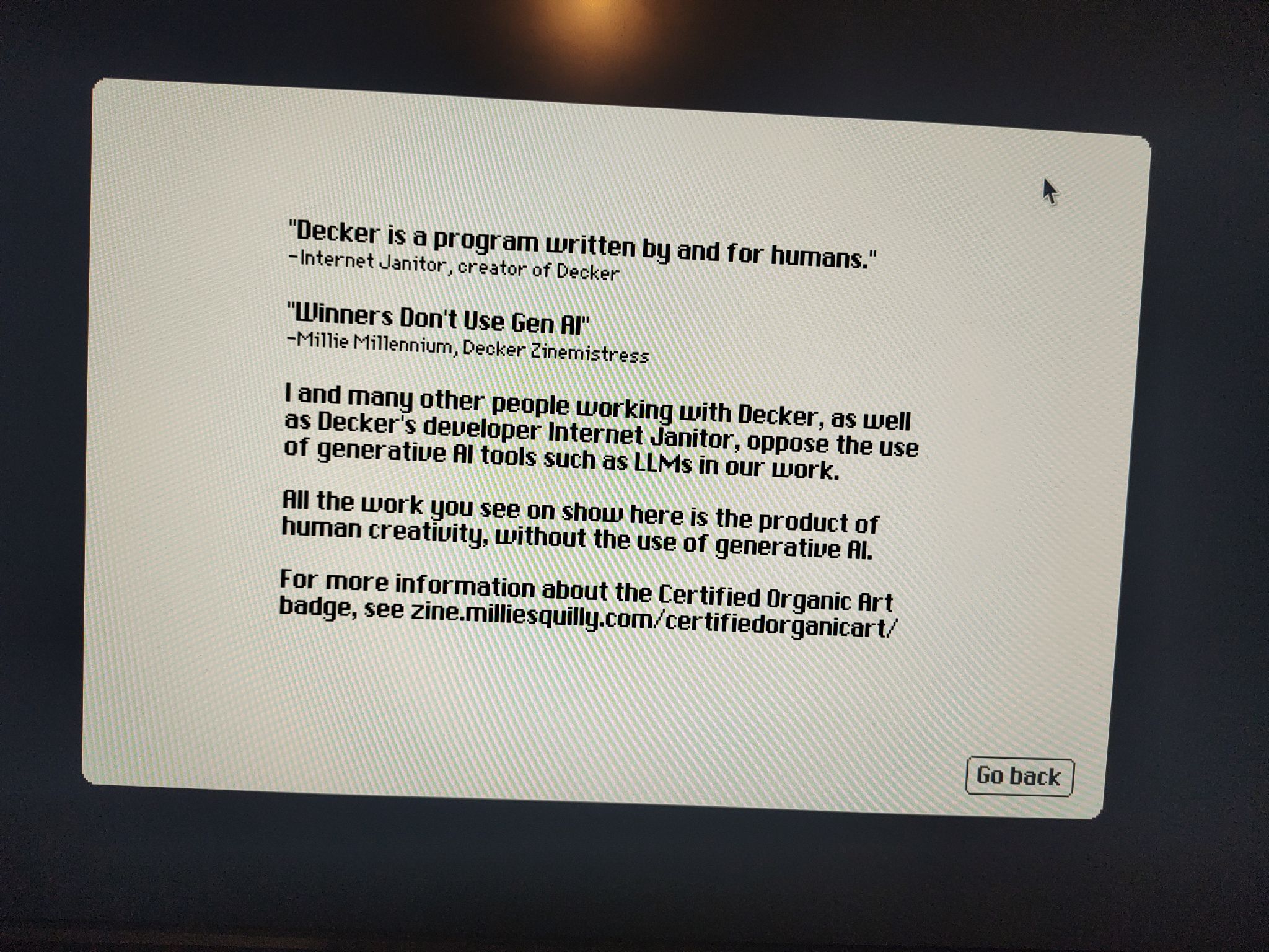 Photo of a screen with the text: "Decker is a program written by and for humans." -Internet Janitor, creator of Decker "Winners Don't Use Gen AI" -Millie Millennium, Decker Zinemistress I and many other people working with Decker, as well as Decker's developer Internet Janitor, oppose the use of generative AI tools such as LLMs in our work. All the work you see on show here is the product of human creativity, without the use of generative AI. For more information about the Certified Organic Art badge, see zine.milliesquilly.com/certifiedorganicart/